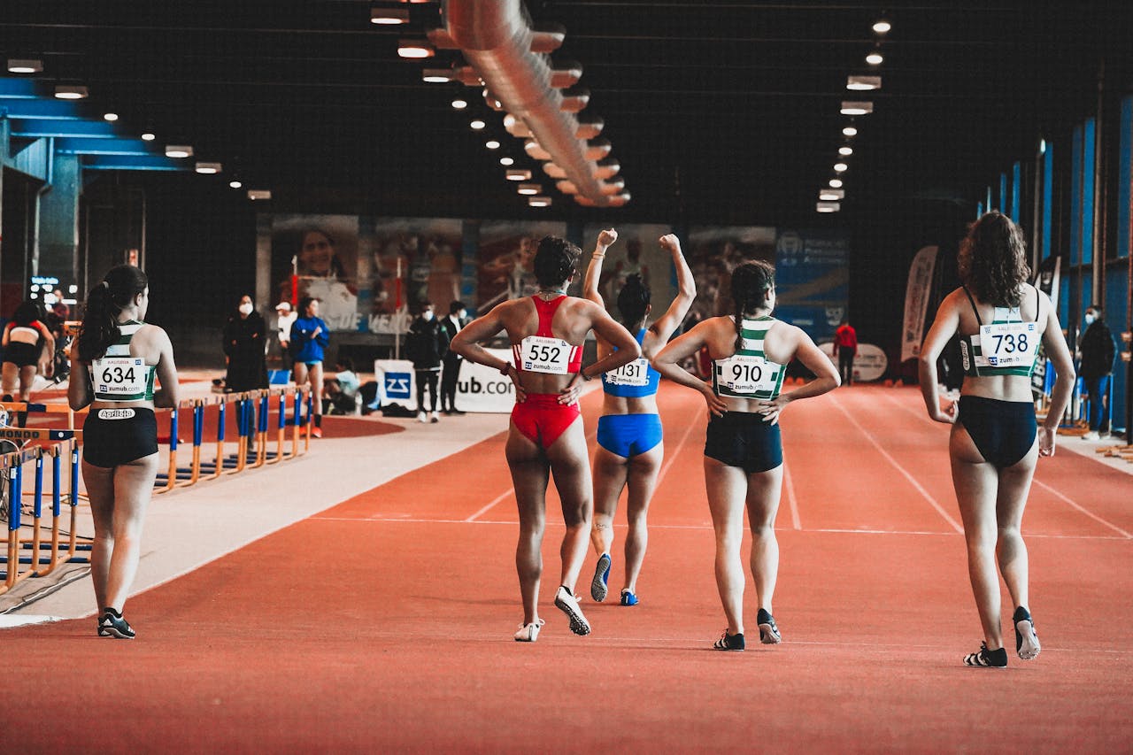 heros-img Group of female athletes at the starting line of an indoor track race, wearing numbered sportswear.